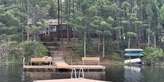 A wooden cabin is nestled among trees by a lake, with a dock extending into the water and a floating platform nearby. Canoes and kayaks are stacked on the shore.