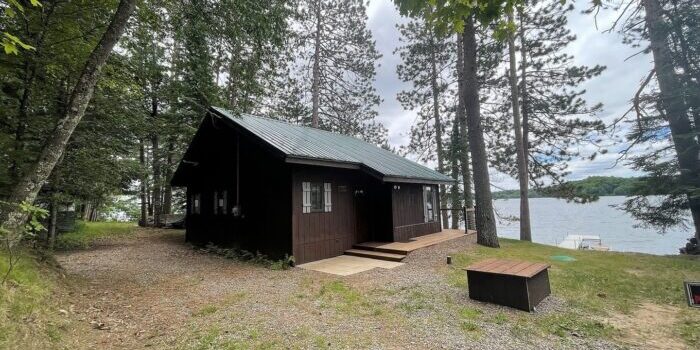 Small dark brown cabin with a green roof surrounded by trees near a lake. A gravel path leads to the cabin, and the lake is visible in the background under a cloudy sky.