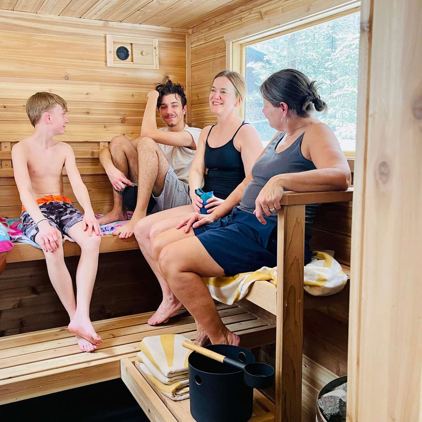 Four people sitting and chatting in a wooden sauna. Two are on the top bench and two are seated below. There is a window in the background showing a snowy scene outside.