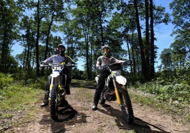 Two motorcyclists wearing full gear and helmets pause on a dirt trail in a forest, with their bikes facing the camera.