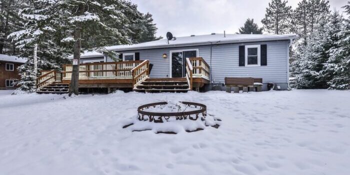 A single-story house with a wooden deck surrounded by snow, featuring a fire pit in the foreground and trees in the background.